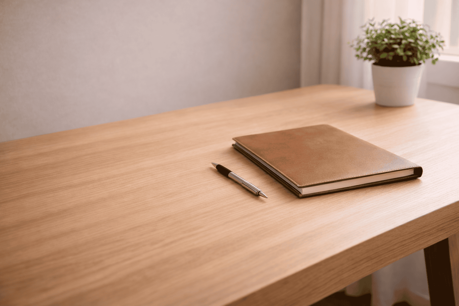A closed brown notebook and a black pen rest on a clean wooden desk. A small potted plant sits in the top right corner near a window with soft natural light.