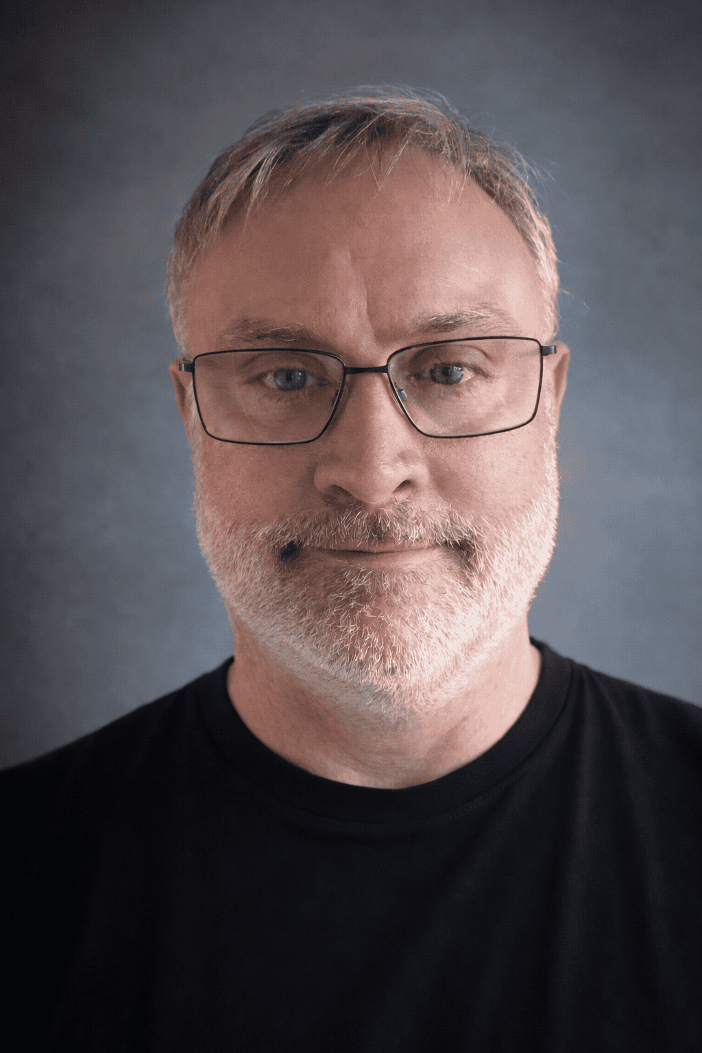 A middle-aged man with short grey hair, glasses, and a beard is wearing a black shirt and looking directly at the camera against a dark, softly blurred background.